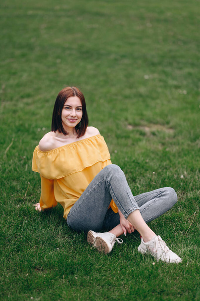 A girl with a square and a yellow blouse sits on a green grass in jeans fell back on her hand, a vertical photo on top - Foto, immagini