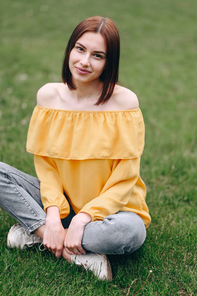 Une fille avec un carré et un chemisier jaune s'assoit sur l'herbe verte et sourit, photo verticale sur le dessus - Photo, image
