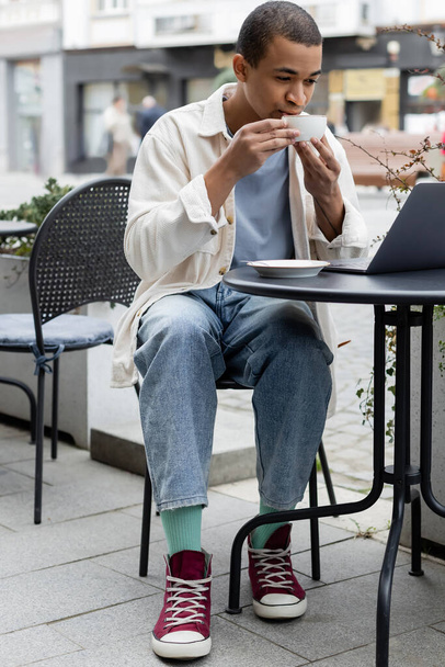 jeune indépendant afro-américain boire du café et en utilisant un ordinateur portable sur la terrasse du café  - Photo, image