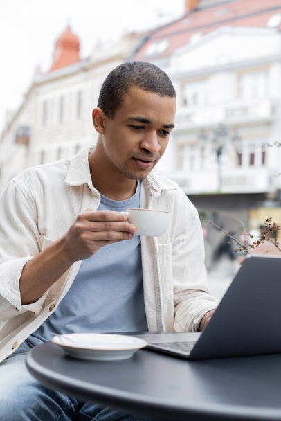 jeune pigiste afro-américain tenant une tasse de café et utilisant un ordinateur portable sur la terrasse du café  - Photo, image