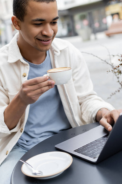 sorridente freelance afroamericano in possesso di una tazza di caffè e utilizzando il computer portatile sulla terrazza del caffè  - Foto, immagini