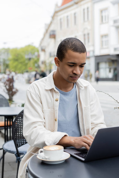 geconcentreerd Afrikaans Amerikaans freelancer met behulp van laptop in de buurt van cappuccino op cafe terras  - Foto, afbeelding
