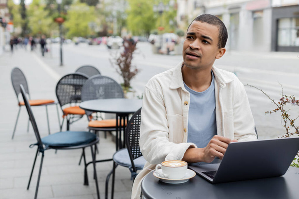 рассеянный африканский американский фрилансер смотрит вдаль возле ноутбука и чашки капучино на террасе кафе  - Фото, изображение