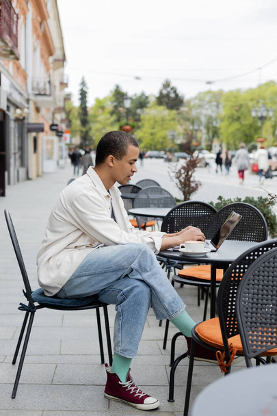 boční pohled na mladý africký americký freelancer pracující na dálku na notebooku v blízkosti šálek cappuccino na kavárně terasa - Fotografie, Obrázek