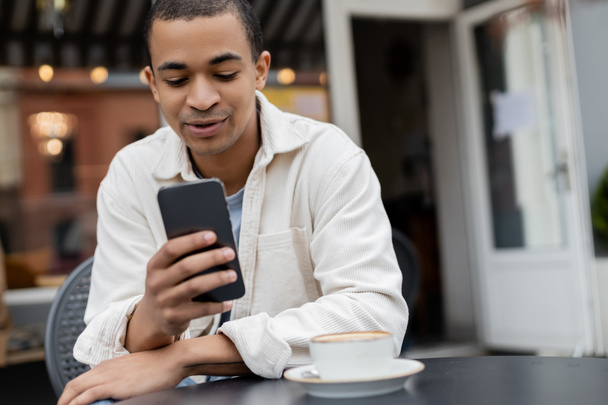 african american man texting on smartphone near cup of cappuccino on table on cafe terrace  - 写真・画像