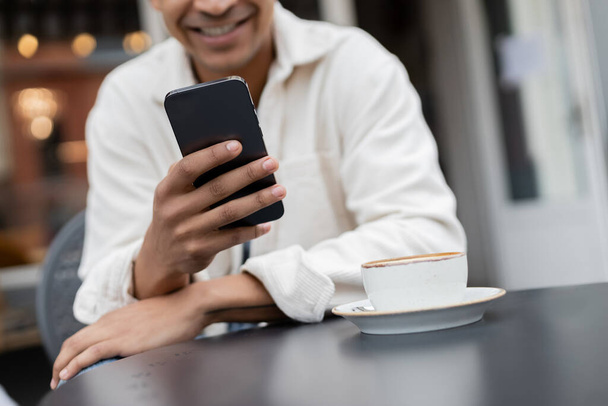 Kesilmiş manzara gülümseyen Afrikalı Amerikalı adam kafenin terasındaki kapuçino bardağının yanında akıllı telefondan mesajlaşıyor.  - Fotoğraf, Görsel