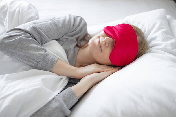 Porträt einer jungen schönen Frau, die während des Schlafs auf dem Bett eine Schlafmaske aus roter Seide trägt. Eine Schlafmaske kann das Licht beim Schlafen blockieren, ermöglicht dem Benutzer einen tieferen Schlaf zu erreichen. - Foto, Bild