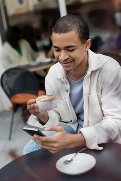 улыбающийся африканский американец пишет смс на смартфоне и держит чашку капучино на террасе кафе  - Фото, изображение