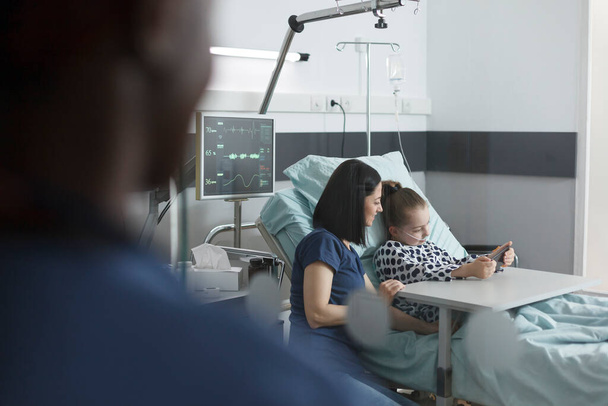 Moeder zitten naast patiënt bed in pediatrische kliniek kamer terwijl schattig klein meisje spelen op smartphone. - Foto, afbeelding
