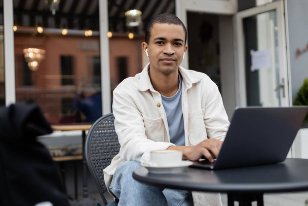 afro-américain pigiste dans des écouteurs sans fil assis près de tasse de café et ordinateur portable sur la terrasse d'été  - Photo, image