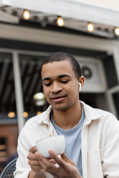 Afrikanisch-amerikanischer Mann mit drahtlosen Kopfhörern blickt auf Tasse Kaffee auf Sommerterrasse  - Foto, Bild