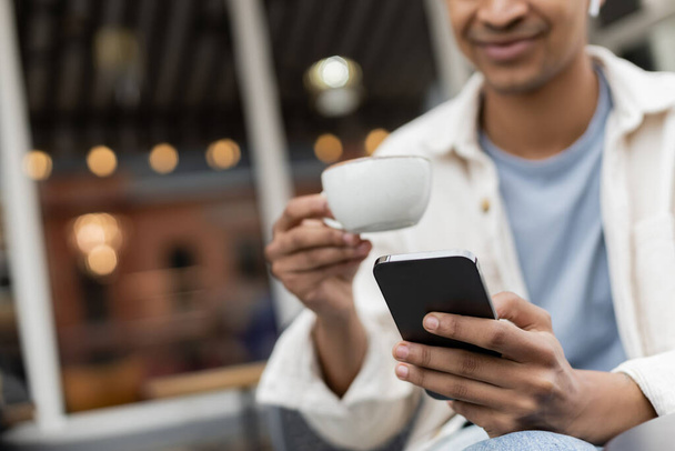 cropped view of happy african american man in wireless earphones using smartphone while holding cup of coffee - Фото, изображение