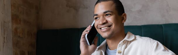 smiling and young african american man talking on smartphone in coffee shop, banner - Photo, Image