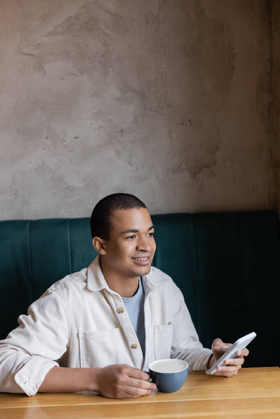 smiling and young african american man holding smartphone and holding cappuccino in coffee shop  - Fotoğraf, Görsel