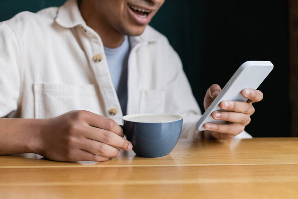 vista recortada de alegre hombre afroamericano mensajes de texto en el teléfono inteligente cerca de la taza en la mesa - Foto, Imagen