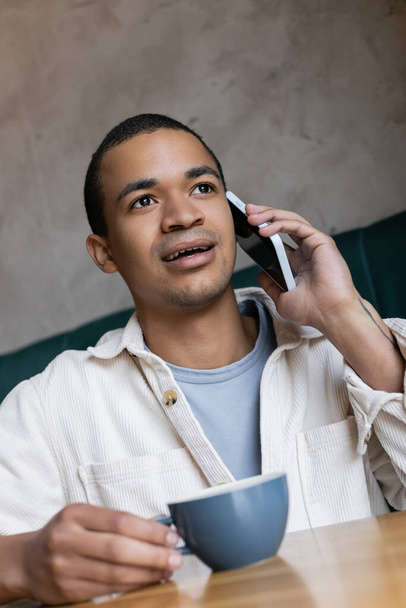 jovem afro-americano homem ter telefonema e segurando xícara de café - Foto, Imagem