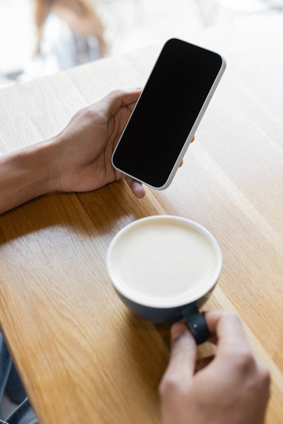 cropped view of blurred african american man holding smartphone with blank screen near cup of cappuccino  - Фото, изображение