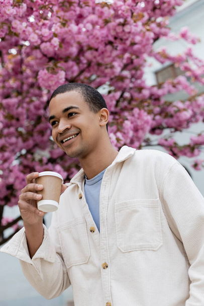 χαμηλή γωνία άποψη του ευτυχισμένος Αφροαμερικανός άνδρας κρατώντας καφέ για να πάει κοντά σε ροζ κερασιά  - Φωτογραφία, εικόνα