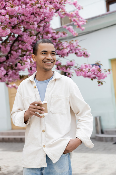 souriant homme afro-américain tenant du café pour aller près de cerisier rose dans la rue - Photo, image