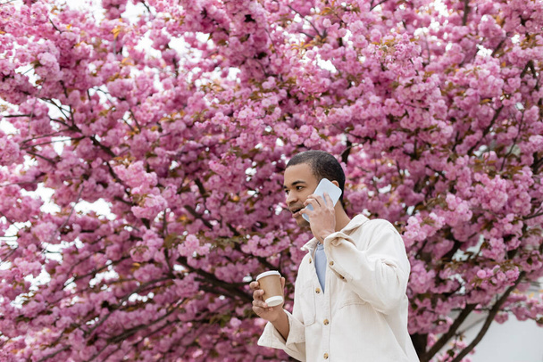 joven afroamericano hombre celebración takeaway beber y hablar en smartphone cerca de rosa cerezo  - Foto, Imagen