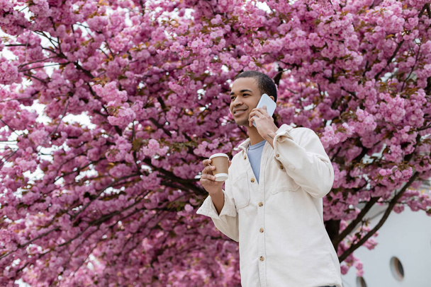 smiling african american man holding takeaway drink and talking on smartphone near pink cherry tree  - Foto, Imagem
