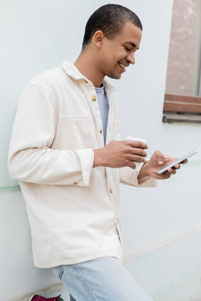 onnellinen afrikkalainen amerikkalainen mies pitää paperimukia ja älypuhelinta lähellä rakennusta kadulla Euroopassa - Valokuva, kuva
