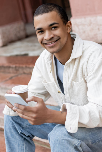 happy african american man holding takeaway beverage and smartphone while sitting on stairs in city  - Foto, immagini
