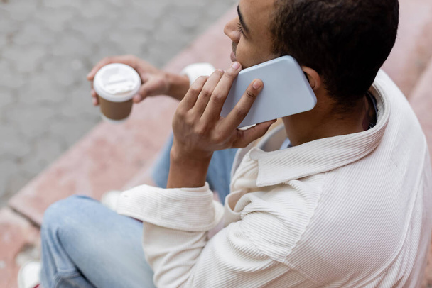 von oben: Afrikanisch-amerikanischer Mann hält Imbissgetränk in der Hand und spricht auf dem Smartphone, während er auf der Treppe in der Stadt sitzt  - Foto, Bild