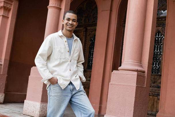 fröhlicher afrikanisch-amerikanischer Mann in Hemdjacke steht mit Händen in Taschen in der Nähe des Gebäudes - Foto, Bild