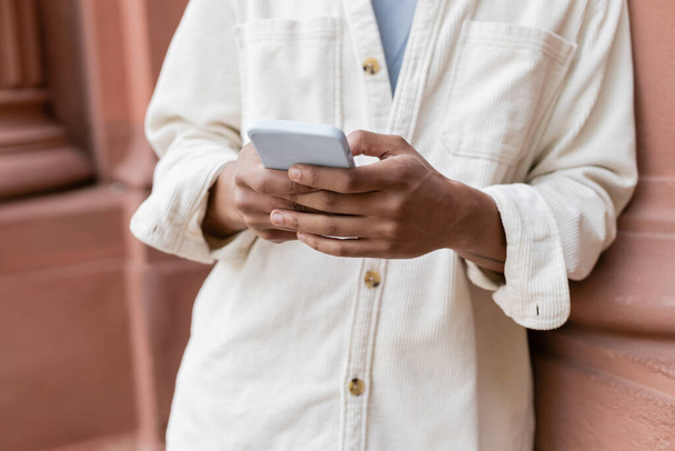cropped view of african american man in shirt jacket messaging on smartphone near building - Photo, image