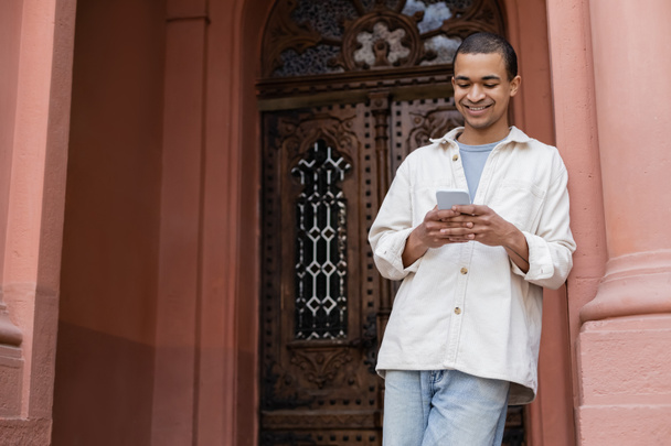 happy african american man in shirt jacket chatting on smartphone near building - Foto, Bild