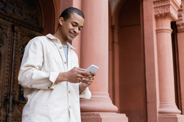 cheerful african american man in stylish shirt jacket messaging on smartphone near building - Photo, image