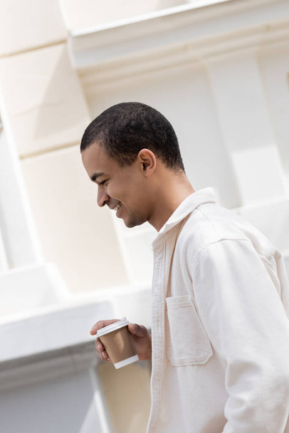 zijaanzicht van gelukkig Afrikaans-Amerikaanse man in shirt jas met koffie te gaan en wandelen in de stad  - Foto, afbeelding