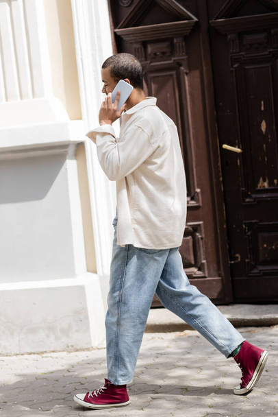 full length of african american man in shirt jacket talking on mobile phone on street in urban city  - Fotografie, Obrázek