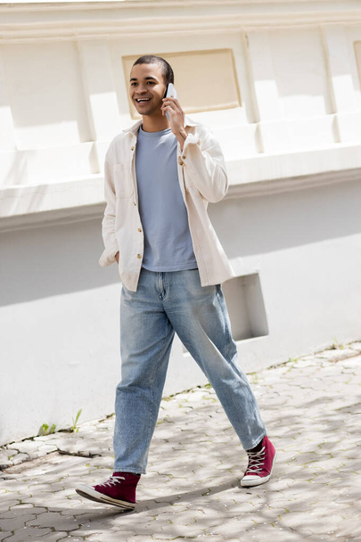 full length of smiling african american man in shirt jacket talking on mobile phone on street in urban city  - Foto, Bild