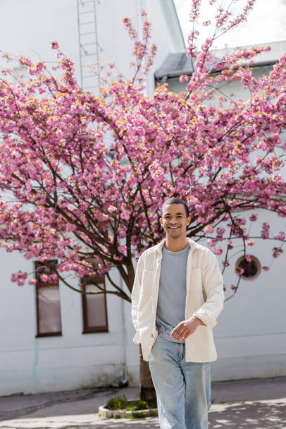 陽気なアフリカ系アメリカ人の男性がシャツのジャケットを着て日本の桜の木の近くのポケットに手を入れて歩く - 写真・画像