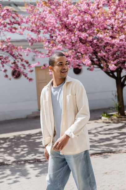 positive african american man in shirt jacket walking near pink cherry tree - Fotoğraf, Görsel