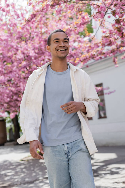 african american man in shirt jacket walking near pink cherry tree - Foto, Bild