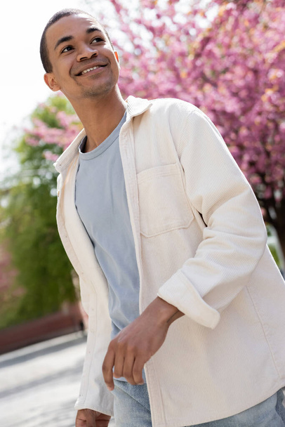 uomo afroamericano in camicia giacca camminare vicino albero di ciliegio rosa - Foto, immagini