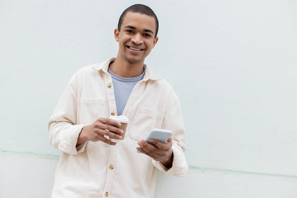 happy african american man smiling while holding smartphone and paper cup outside - Photo, Image