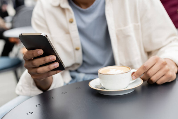 partial view of african american man texting on smartphone near cappuccino on table - Photo, Image