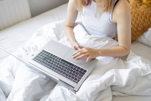 ベッドの上に座っているパジャマ姿の若い女性の作物のショットは、午前中にラップトップを使用しています。家庭で働くこと、フリーランスのコンセプト. - 写真・画像