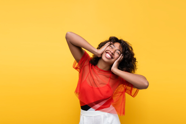 happy african american woman touching face while smiling with closed eyes isolated on yellow - Photo, image