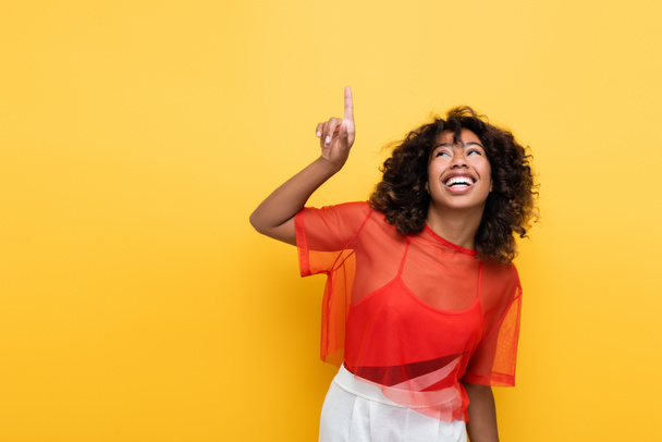 smiling african american woman in summer clothes looking up and pointing with finger isolated on yellow - Foto, Imagem