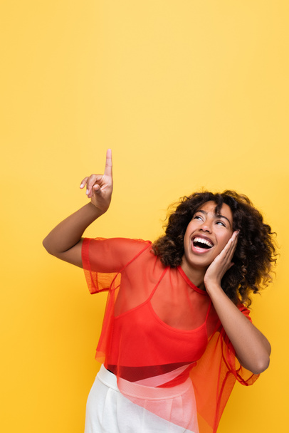 excited african american woman in red top looking up and pointing with finger isolated on yellow - Фото, изображение