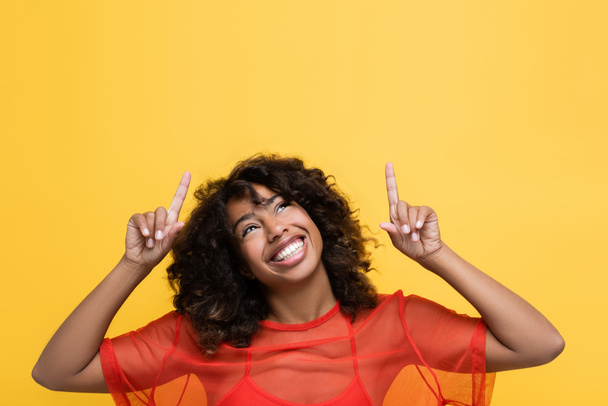 glimlachende Afro-Amerikaanse vrouw omhoog kijken en wijzen met de vingers geïsoleerd op geel - Foto, afbeelding