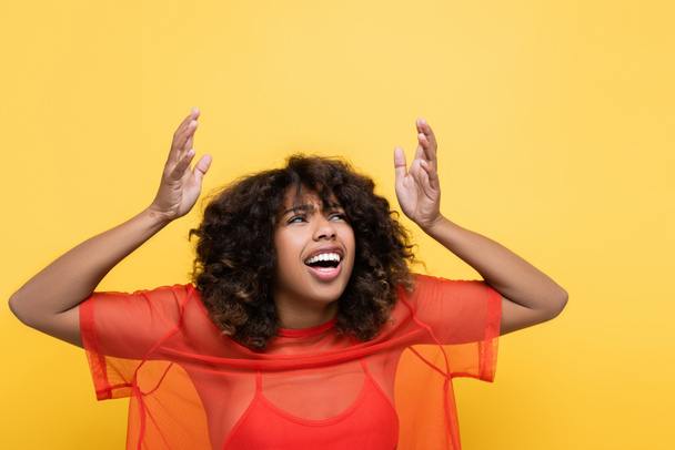 donna afroamericana scioccata in abiti rossi guardando in alto e indicando con le mani isolate sul giallo - Foto, immagini