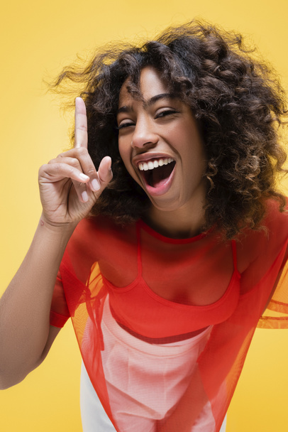 femme afro-américaine étonnée regardant la caméra et montrant le signe d'idée isolé sur jaune - Photo, image