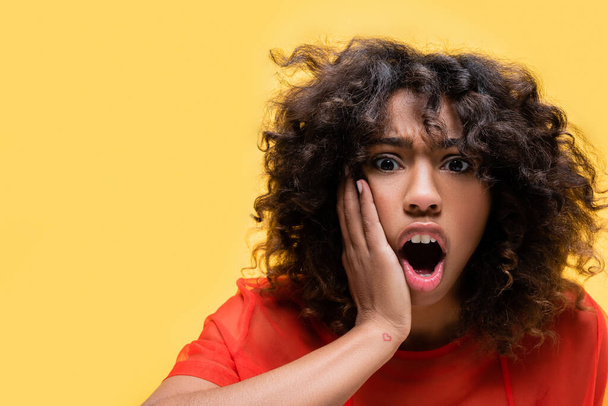 shocked african american woman with open mouth touching face isolated on yellow - Фото, изображение