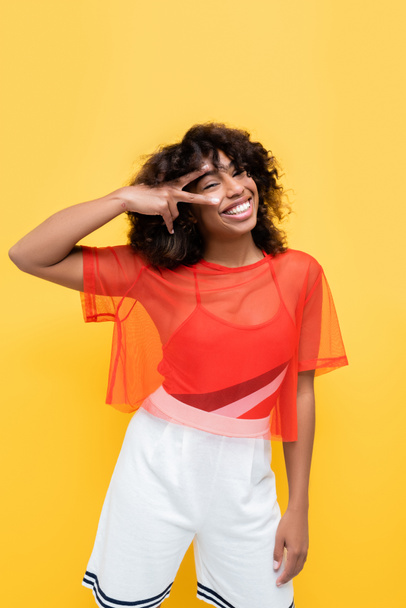 happy african american woman in summer outfit showing victory gesture near eye isolated on yellow - Photo, Image